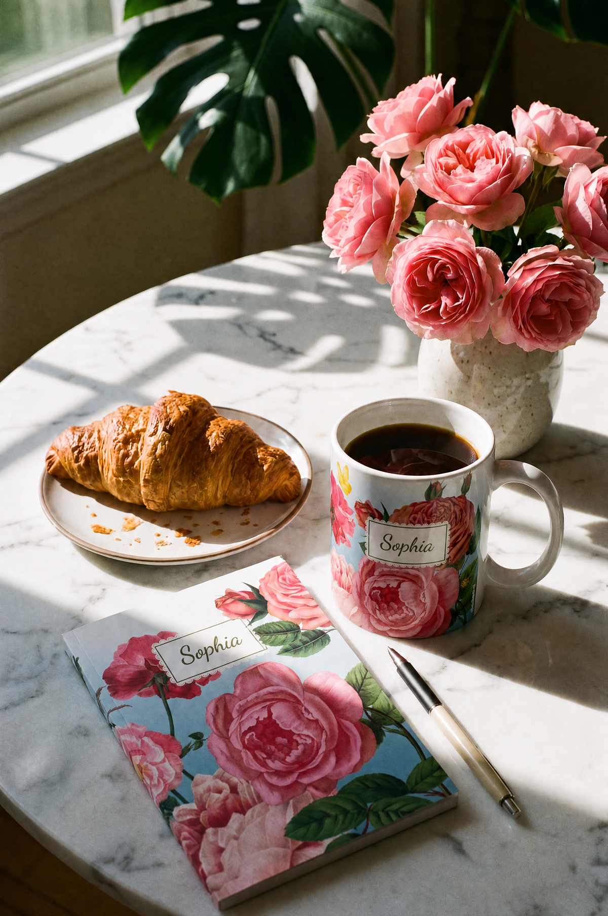 A styled breakfast scene on a marble table with a floral rose garden personalised notebook, a coffee mug, a croissant on a plate, and a vase of pink roses under natural window light.