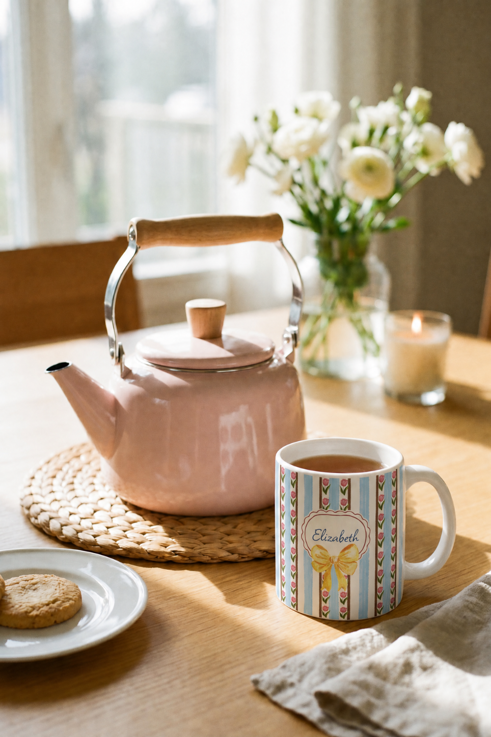 A pink enamel tea kettle with a wooden handle sits on a woven placemat on a wooden table, next to a white ceramic mug with colorful stripes and a personalized name label reading 'Elizabeth'. A small plate with a cookie, a candle, and a vase of white flowers are arranged in a warm, sunlit room.