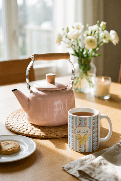 A pink enamel tea kettle with a wooden handle sits on a woven placemat on a wooden table, next to a white ceramic mug with colorful stripes and a personalized name label reading 'Elizabeth'. A small plate with a cookie, a candle, and a vase of white flowers are arranged in a warm, sunlit room.