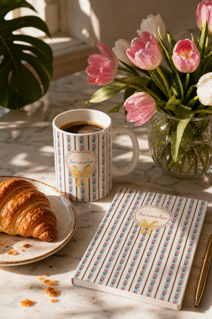 A styled breakfast scene on a marble table with a personalised ceramic tulip mug filled with coffee, a croissant on a plate, and a matching patterned notebook in front. A glass vase of pink tulips sits in the background.