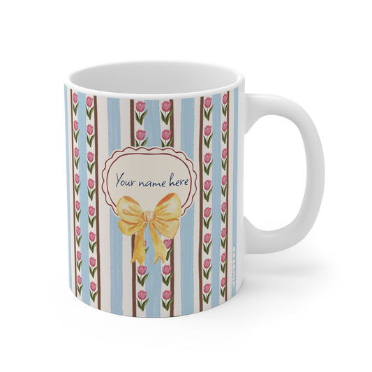 A white ceramic mug with a decorative vertical stripe pattern featuring light blue, brown, and white stripes, pink tulip flowers, and a central cream label with a yellow bow and the text 'Your name here'.