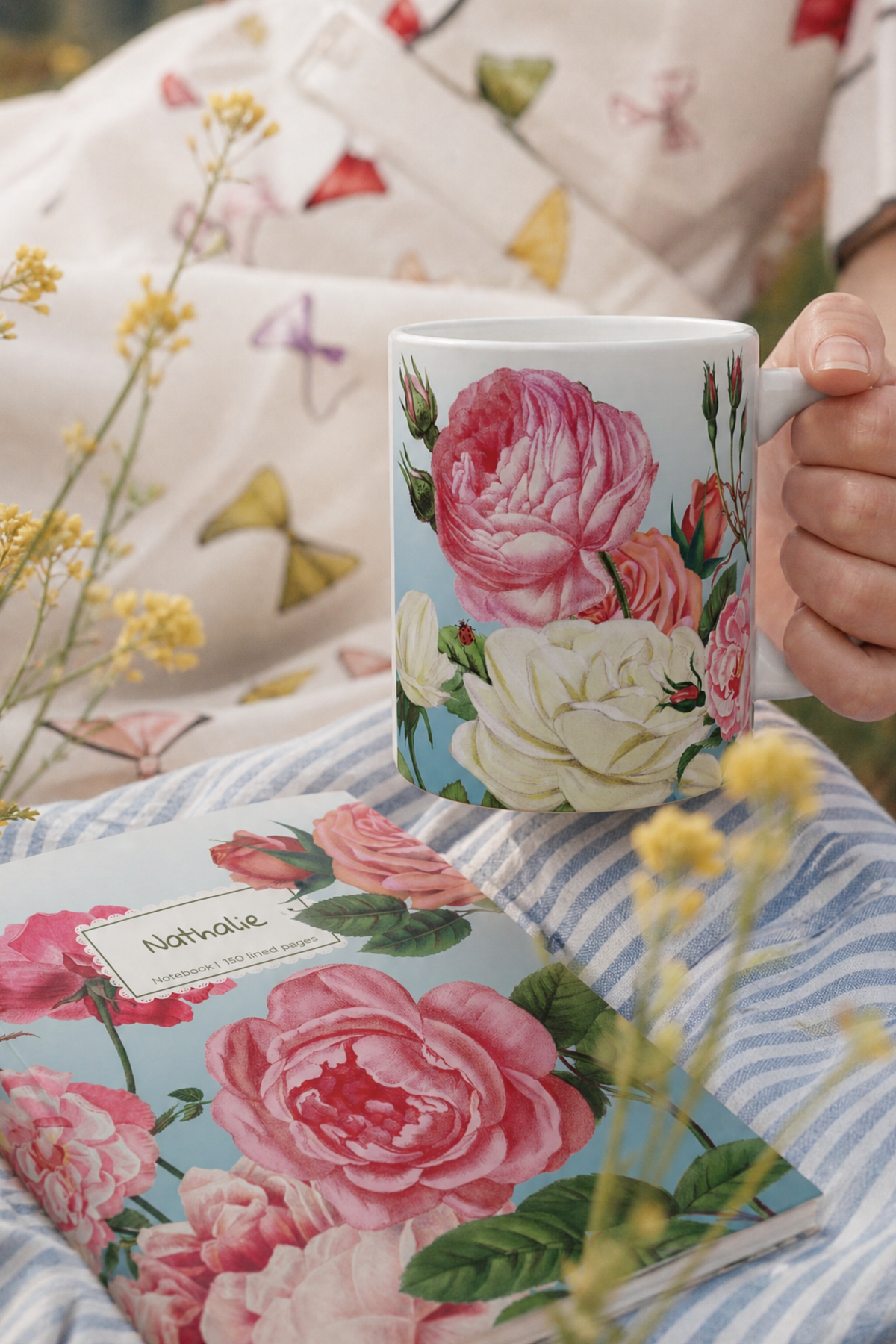 A white ceramic mug with a pink and cream floral print of a rose garden is held by a hand, set against a cozy background of a matching floral blanket and a blue-and-white striped pillow. A matching floral notebook lies in the foreground with a personalised label.